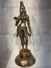 A tall, elegant brass statue of the Hindu goddess Lakshmi, standing gracefully with intricate details on her clothing and adornments, set against a subtly textured, light-colored wall.